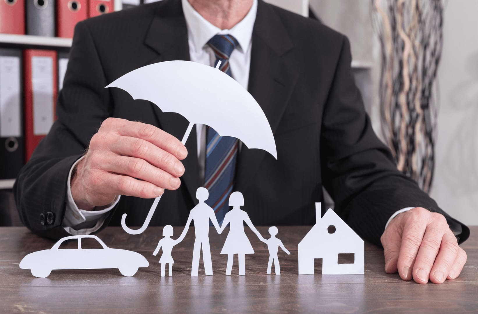 un homme tient un parapluie en papier au dessus d'une voiture, d'une maison et d'une famille qui sont également en papier; le parapluie représente la protection relative que confère le fonds de garantie des assurances aux assurés