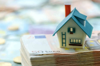 Maison miniature posée sur un tas de billets pour illustrer article sur les arnaques à la location saisonnière