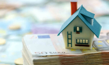 Maison miniature posée sur un tas de billets pour illustrer article sur les arnaques à la location saisonnière